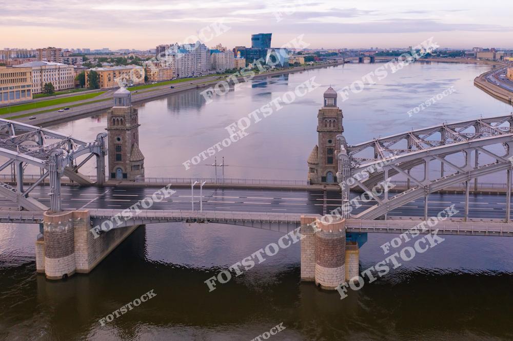 На фотографии изображен Большеохтинский мост в Санкт-Петербурге, пересекающий реку Неву. Этот мост выделяется своими массивными башнями и металлическими арками. На заднем плане видны просторные набережные и современные здания, отражающие гармонию исторической и современной архитектуры города. Тихая атмосфера утра подчеркивает величие моста.